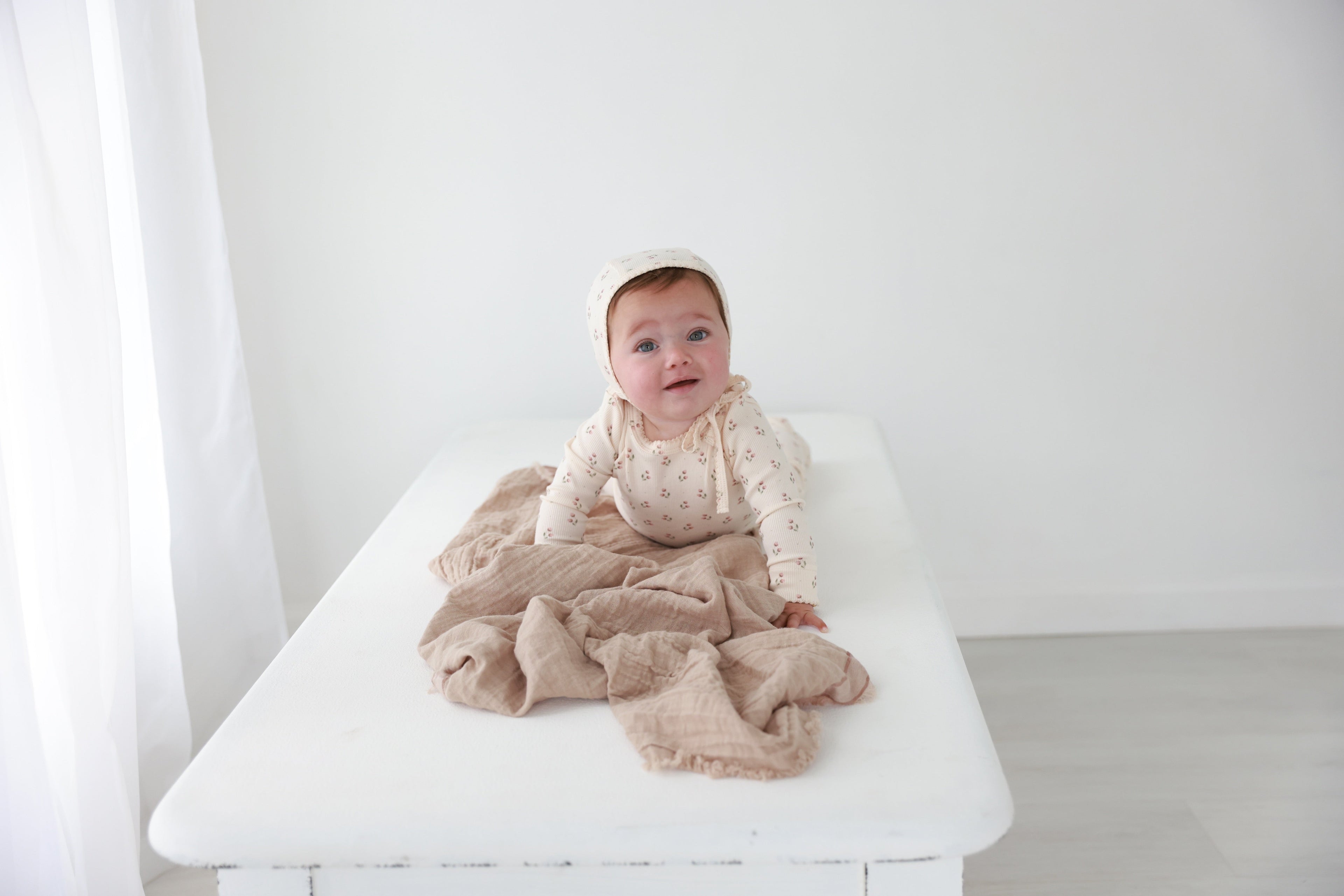 Tiny Tulip layette set for girls by Adee, baby in cream floral bonnet and matching bodysuit on a white bed.