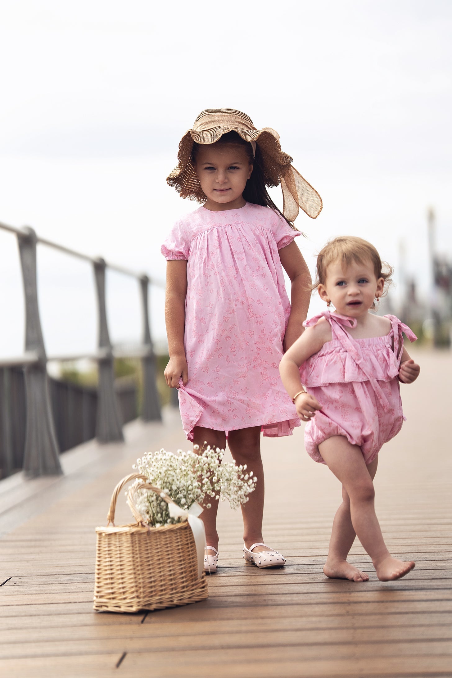 Two young girls in pink flower dresses with short sleeves on a boardwalk; one wears a straw hat.