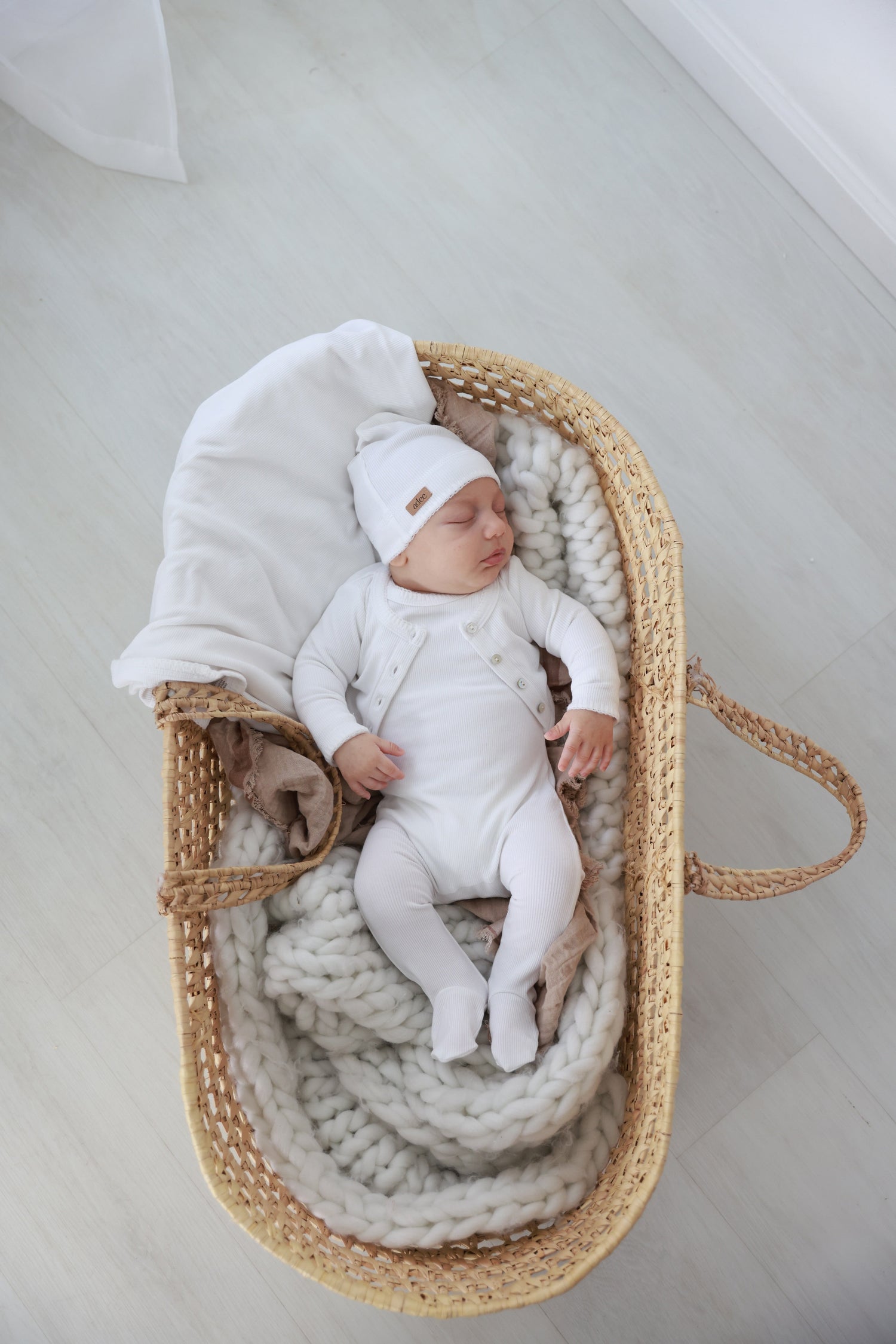 Legacy Stitch White layette set by Adee; infant in white bodysuit and hat in a wicker basket with chunky knit blanket.