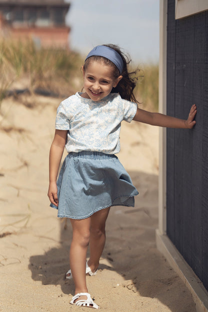 Girl wearing a blue flowered short sleeve T-shirt with a light blue skirt, smiling on the beach.