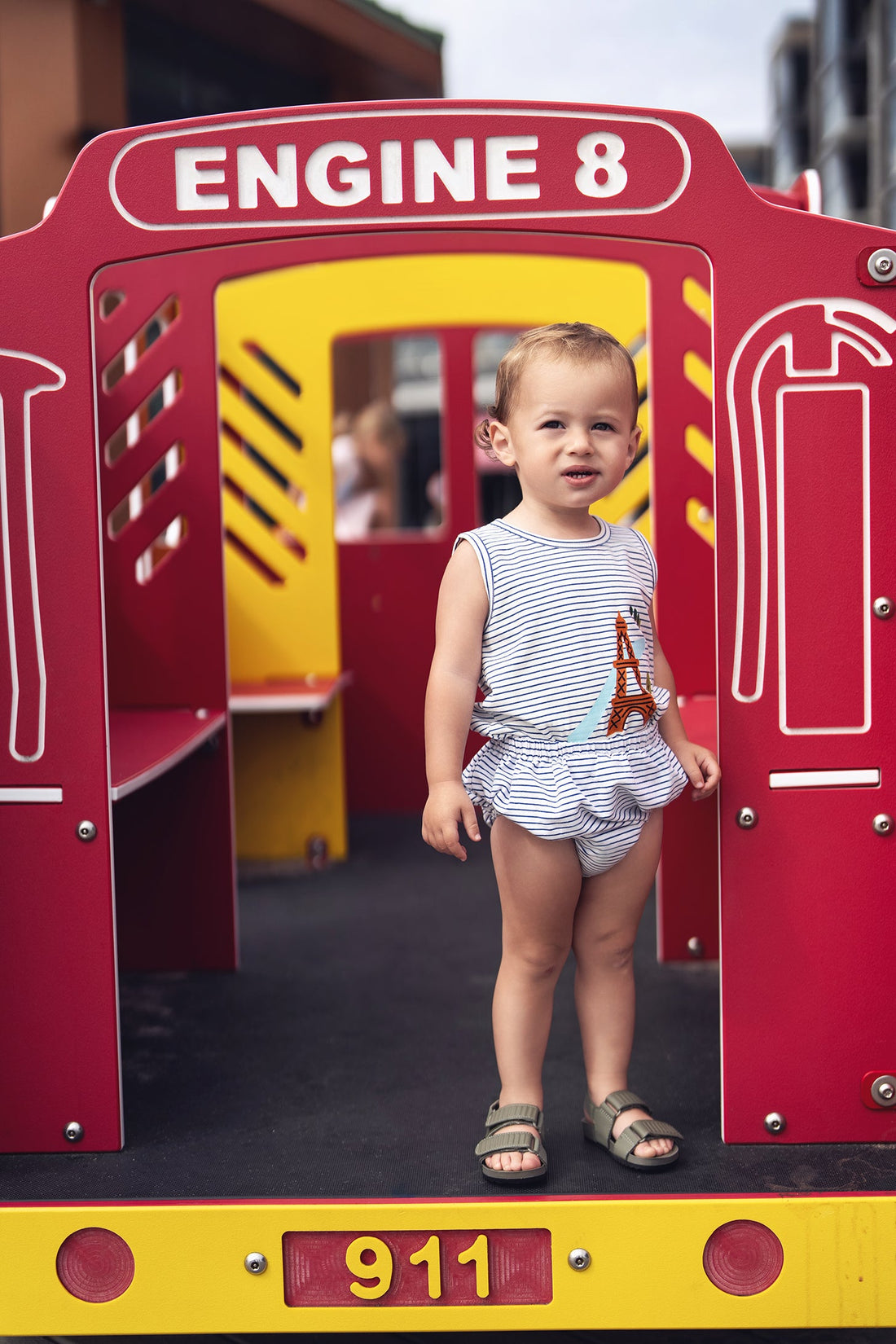 Toddler in Eiffel Tower bloomers & striped tank set, standing at a colorful playground.